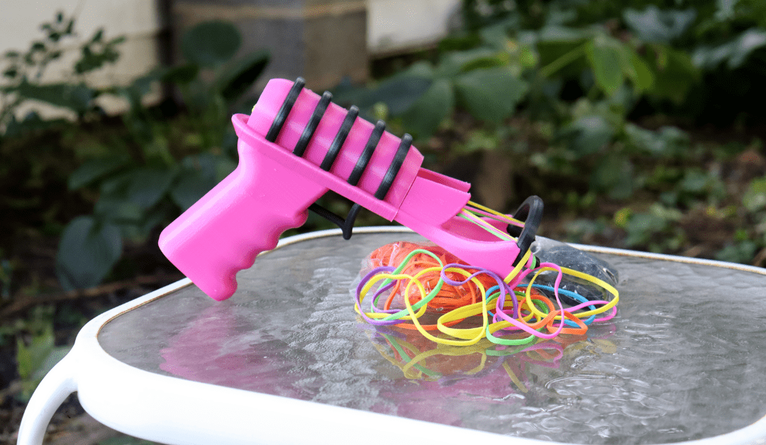 The Snapper 2.0 sitting on a table. The product is a 3-D printed hot pink and black ray gun-looking design, and it's surrounded by rainbow rubber bands.