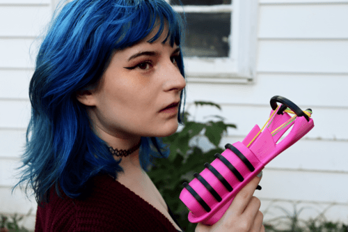 A woman with blue hair and winged eyeliner holds Terrible Toyshop's The Snapper 2.0 with one finger on the trigger. She looks dramatically into the distance. 